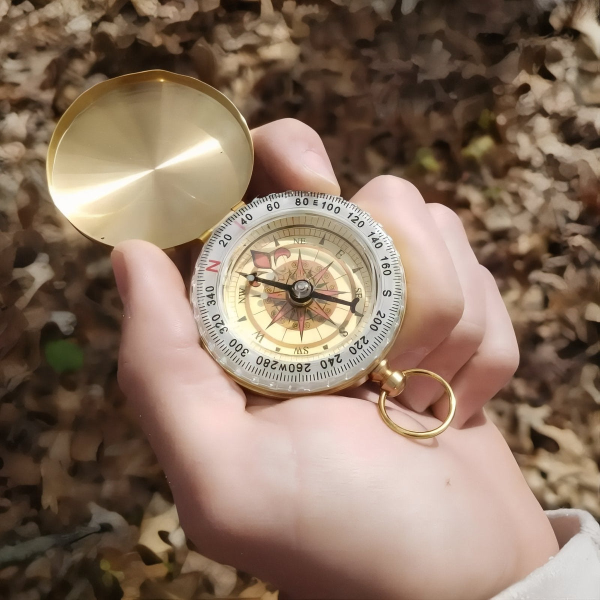 Open gold-tone pocket compass held in a palm over autumn leaves