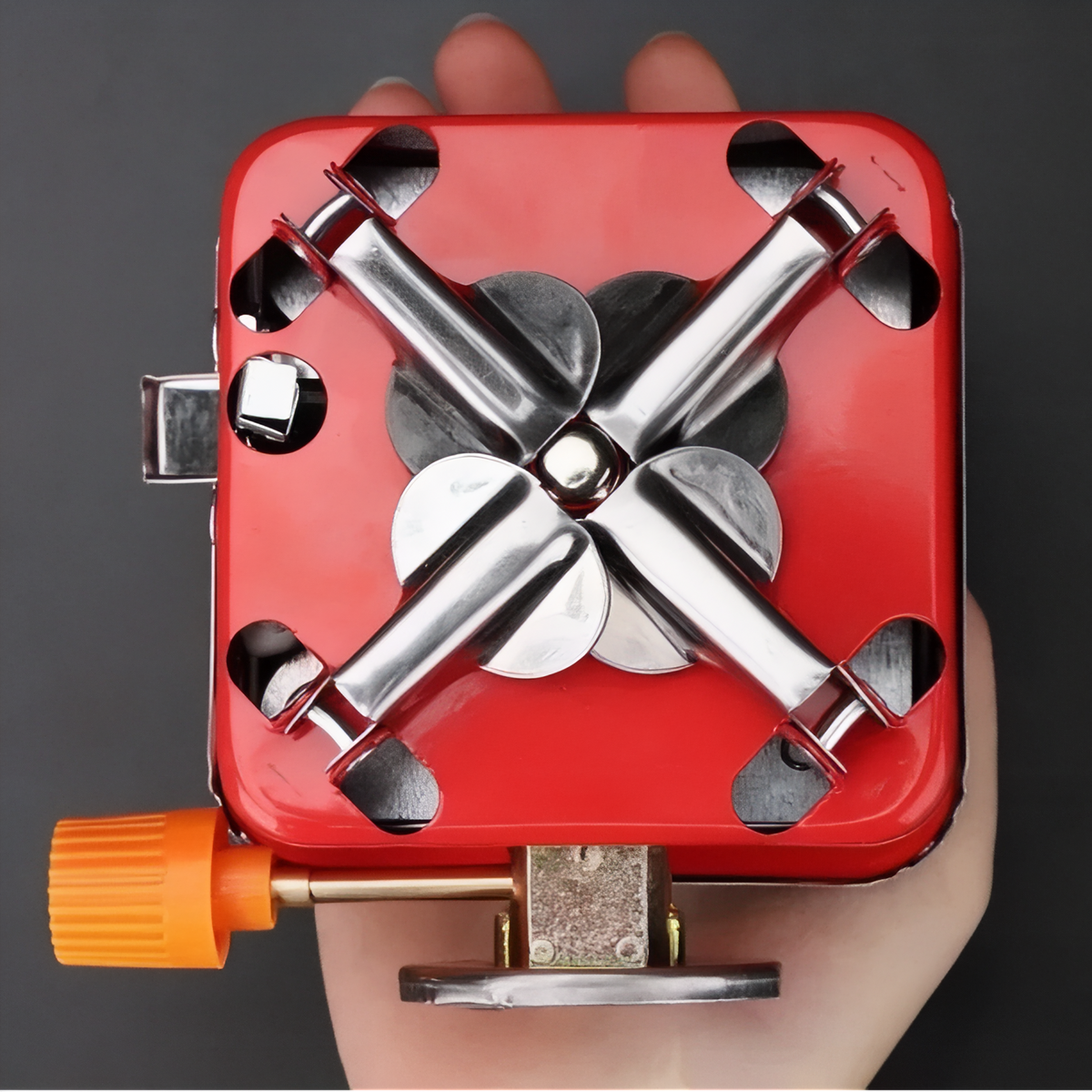 Underside of the folded red stove held in a hand, legs and supports folded in, orange knob at the side
