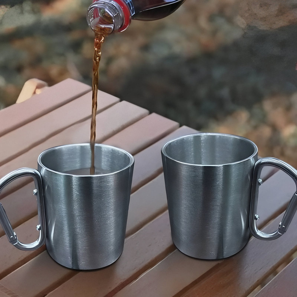Coffee being poured into one of two stainless steel carabiner mugs on a wooden camp table