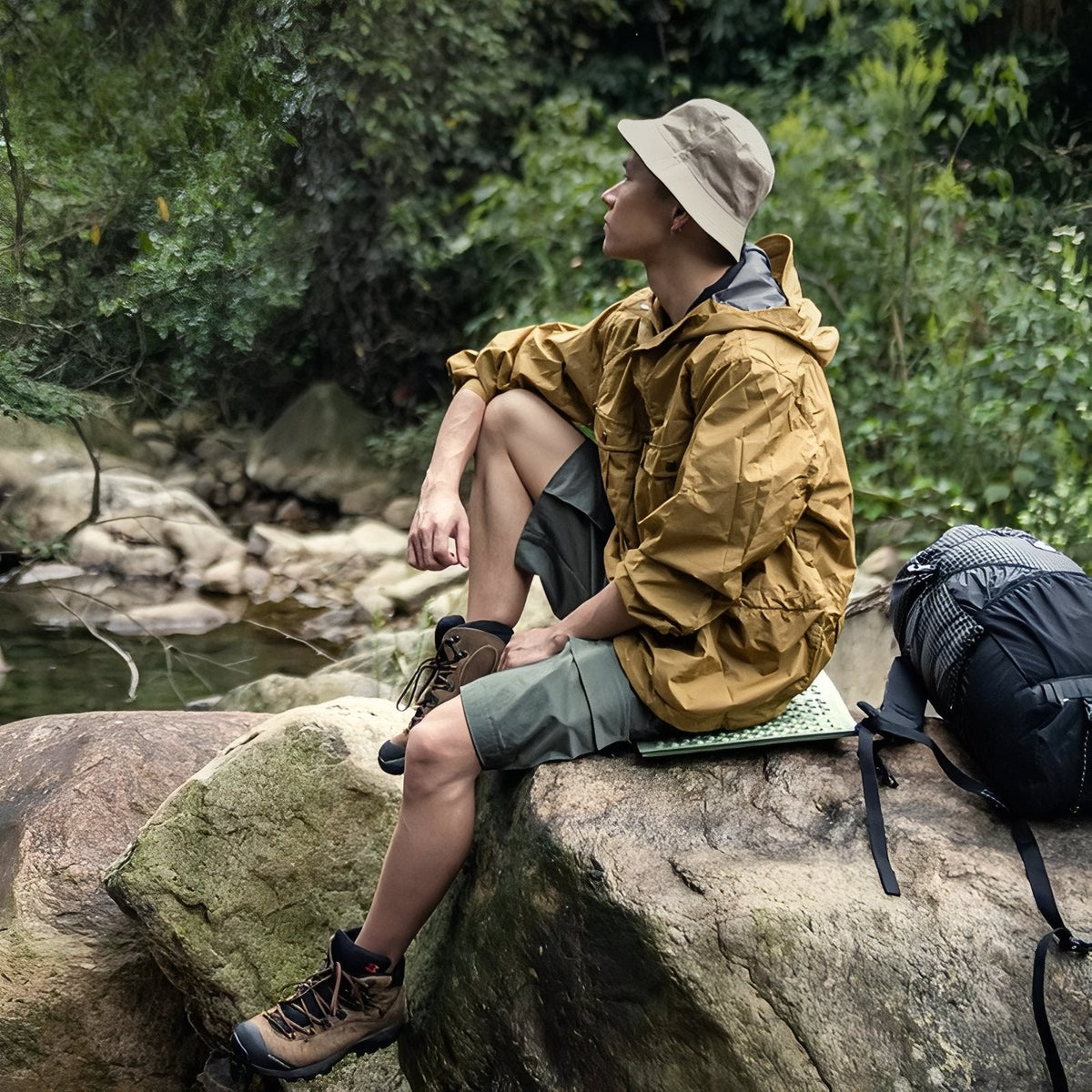 A hiker in a sun hat sitting on the green foam sit pad on a large rock beside a stream, a backpack next to him