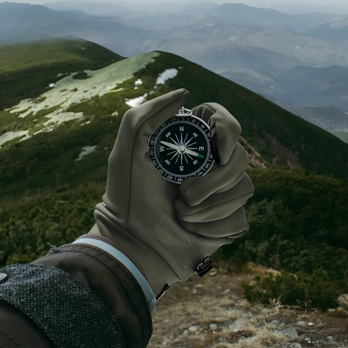 Hand in an army green stretch glove holding a compass above a green mountain valley
