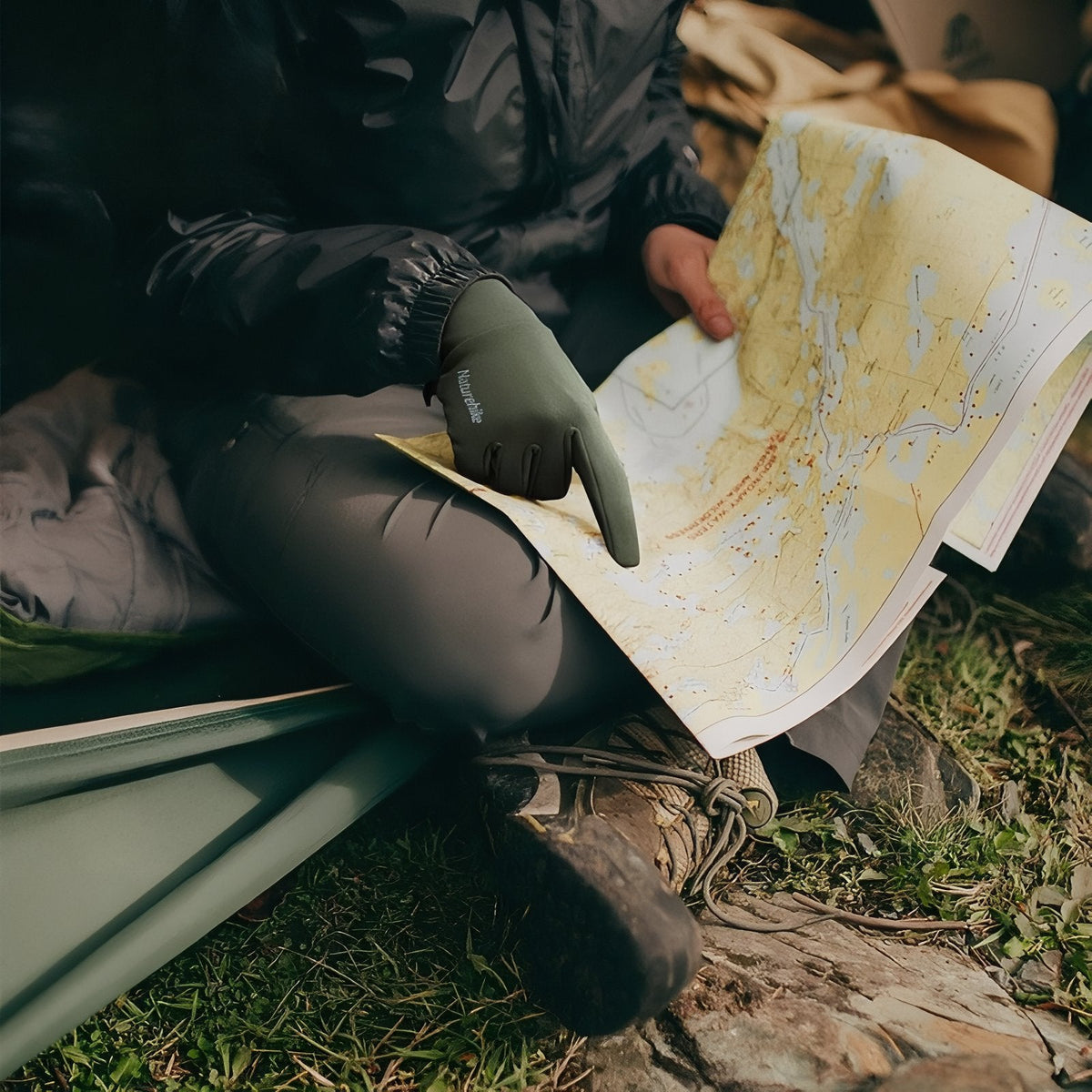 Hiker sitting at a tent reading a paper map, pointing with an army green stretch glove