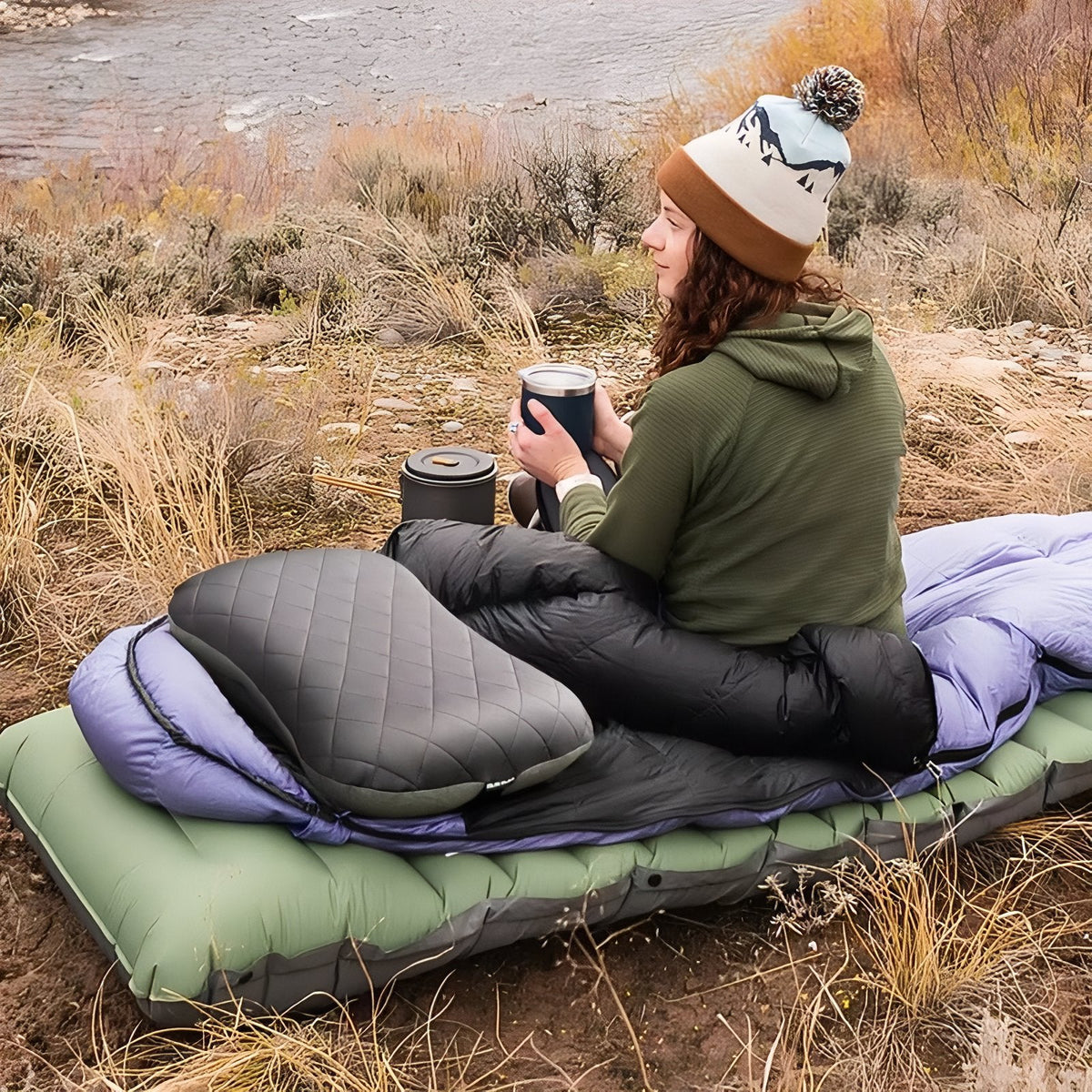 Woman in a sleeping bag sitting on the dark green mat outdoors with a hot drink
