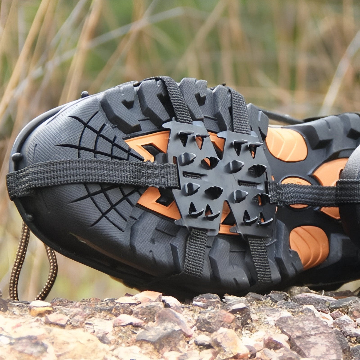 Crampon stretched over a hiking boot sole resting on rocky ground, showing the spike plate and the rubber harness straps