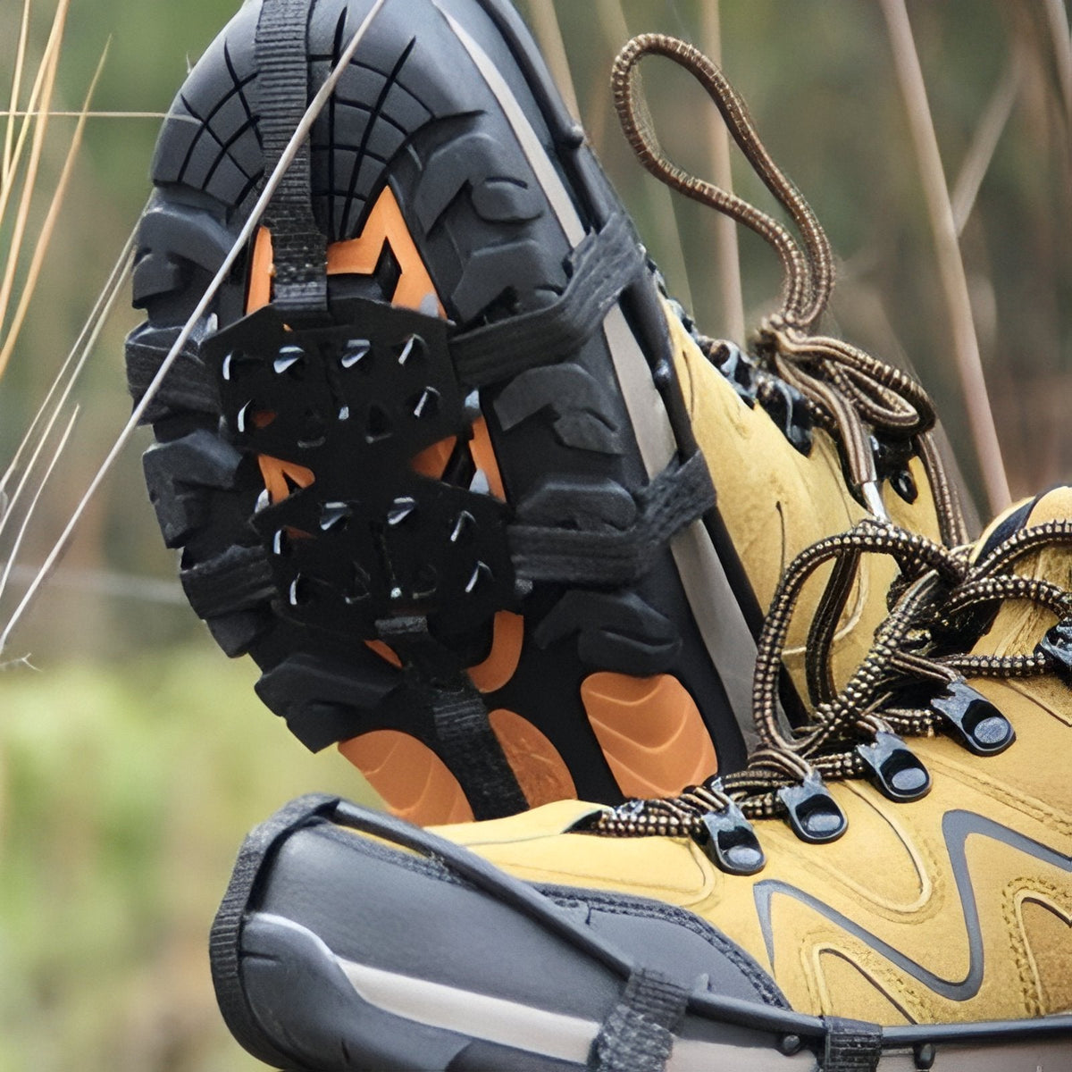 Close-up of a boot sole with the crampon's double forefoot spike plate and rubber straps, outdoors among dry grass