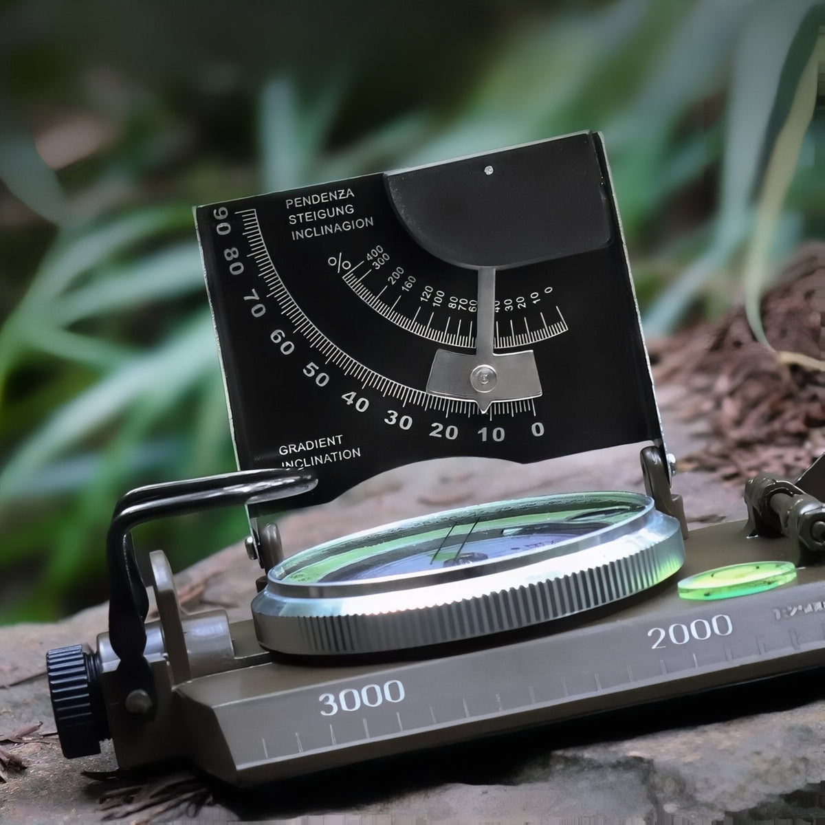 Open sighting compass resting on a mossy rock with its clinometer lid raised and the metal map scale marked 2000 and 3000