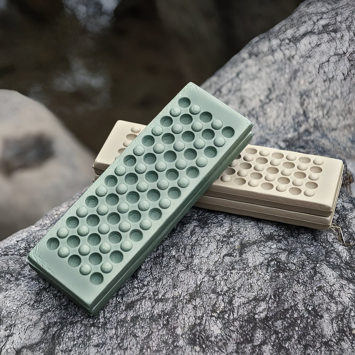A folded sage green sit pad lying on a folded khaki one on a grey rock outdoors