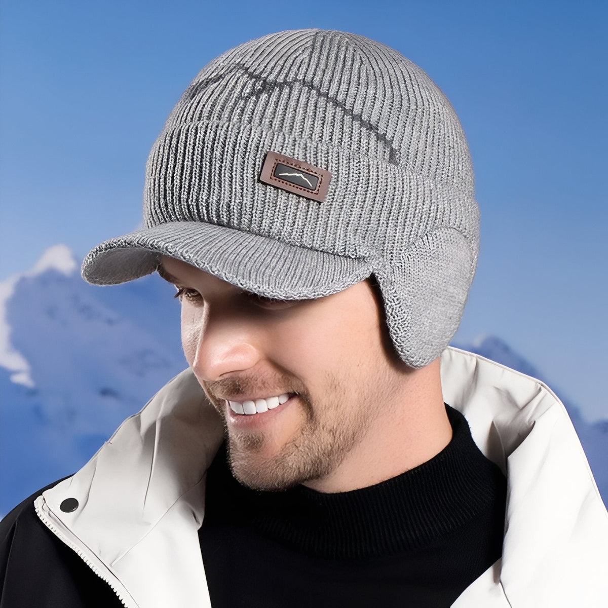 Man wearing the light grey knitted peaked beanie with ear flaps, snowy mountains behind