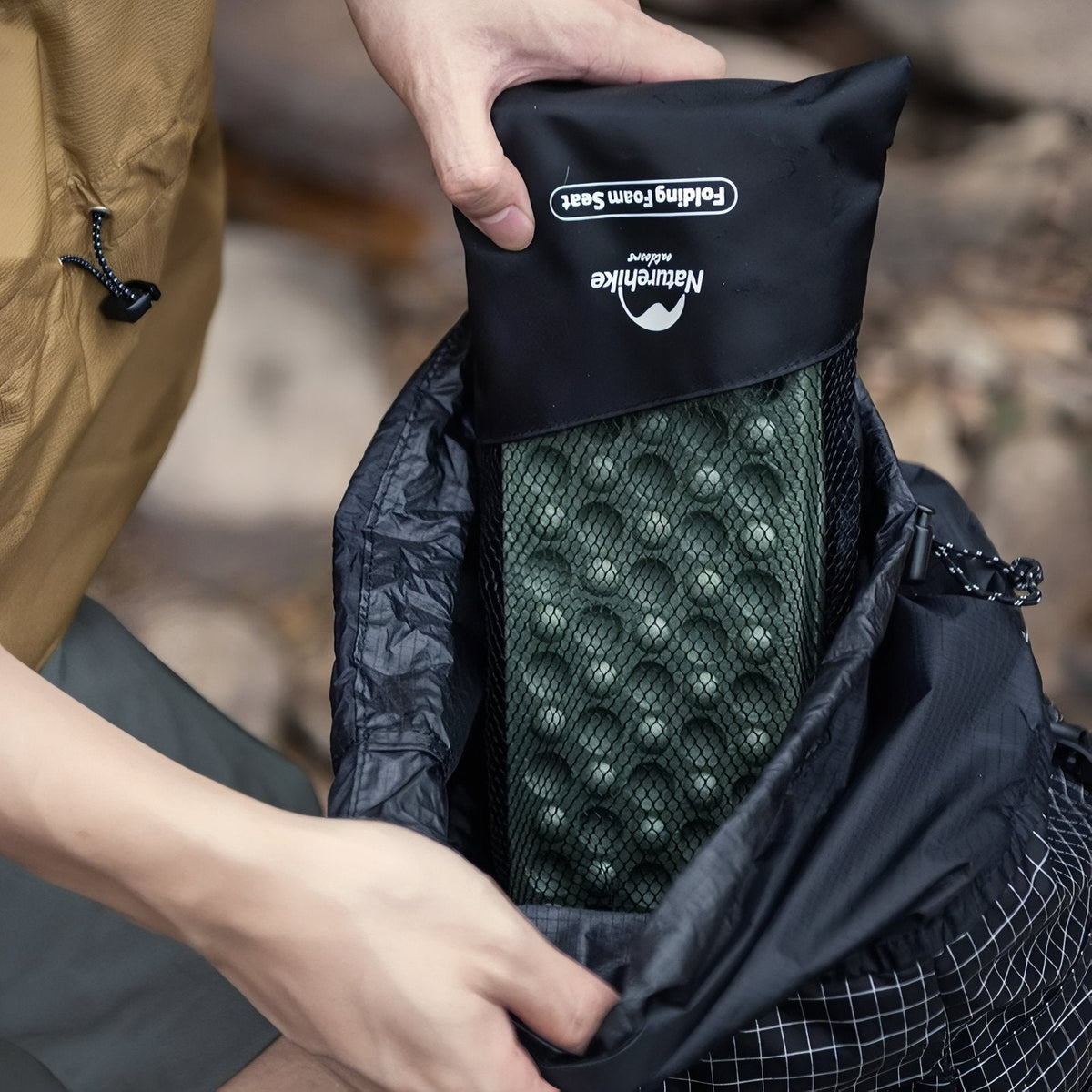 Hands sliding the folded green sit pad in its black storage bag with a mesh window into a backpack