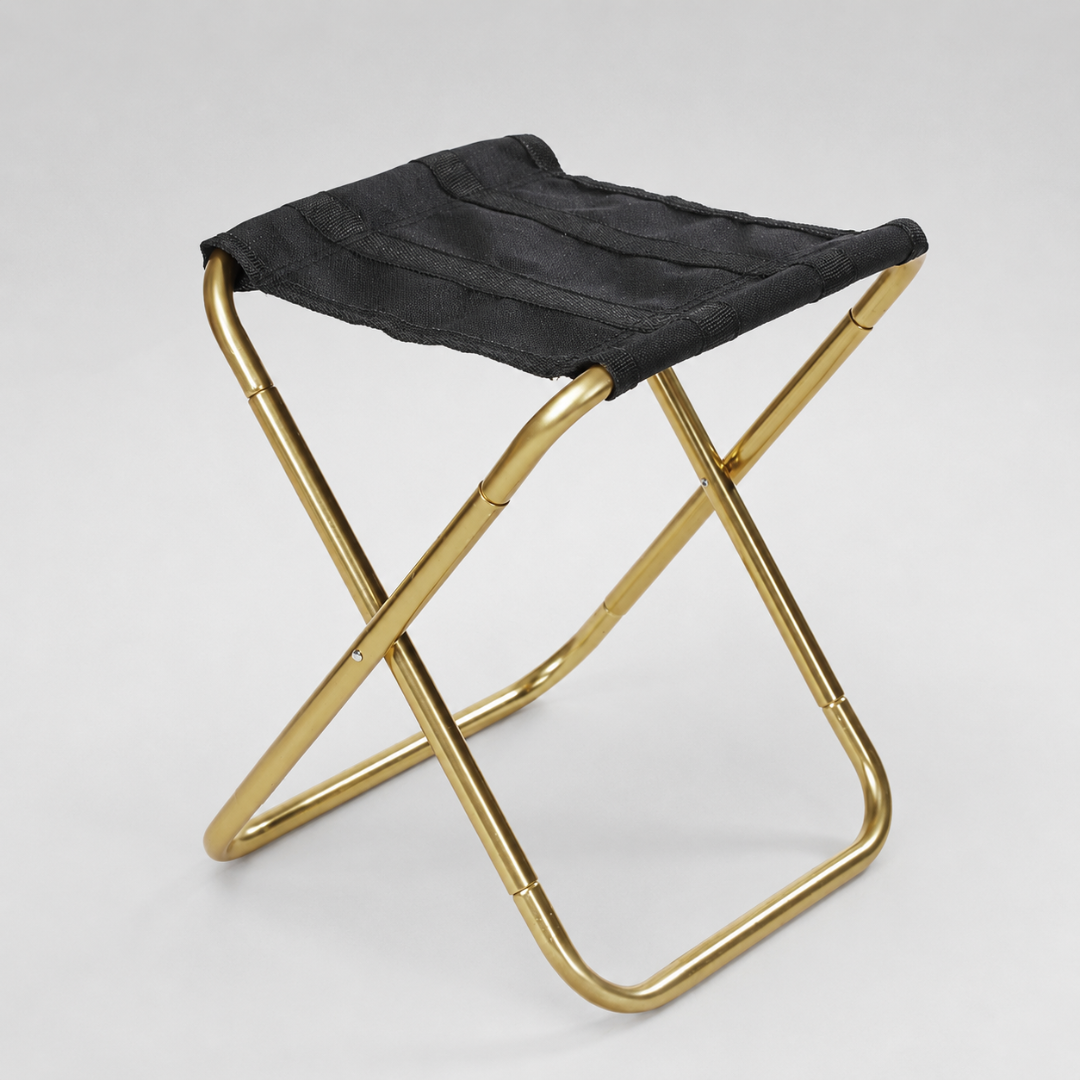 A gold-framed folding stool with a black canvas seat, shown at a three-quarter angle against a plain grey background.
