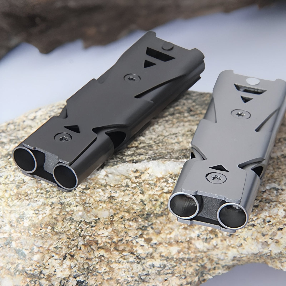 Matt black and silver grey double-tube whistles on a granite stone, showing the two tube openings