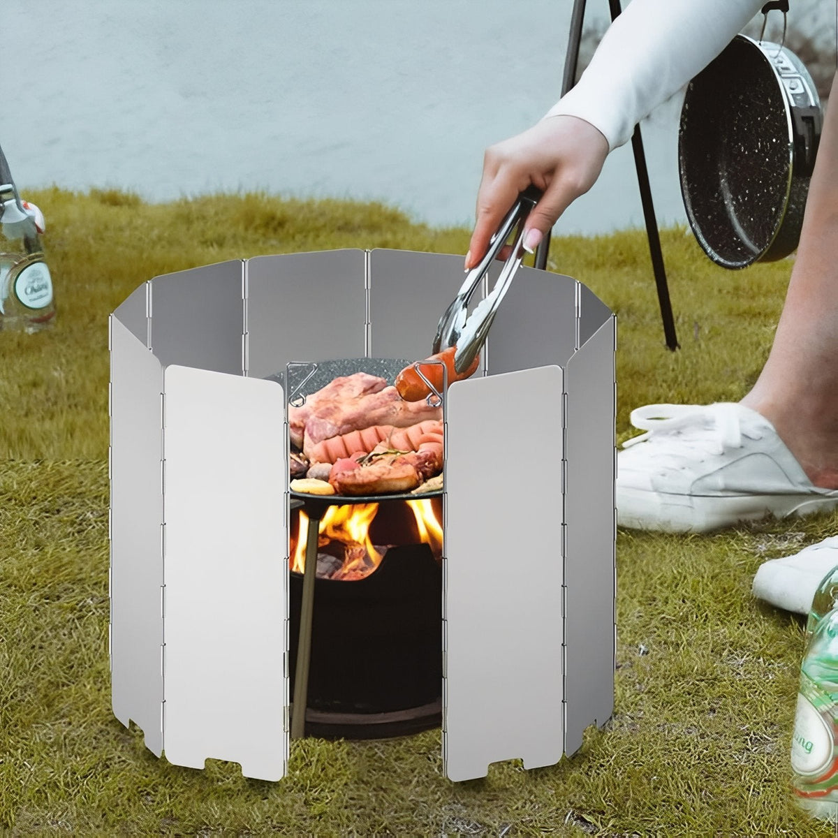 Aluminium windscreen round a small charcoal grill on the grass, a hand turning sausages with tongs