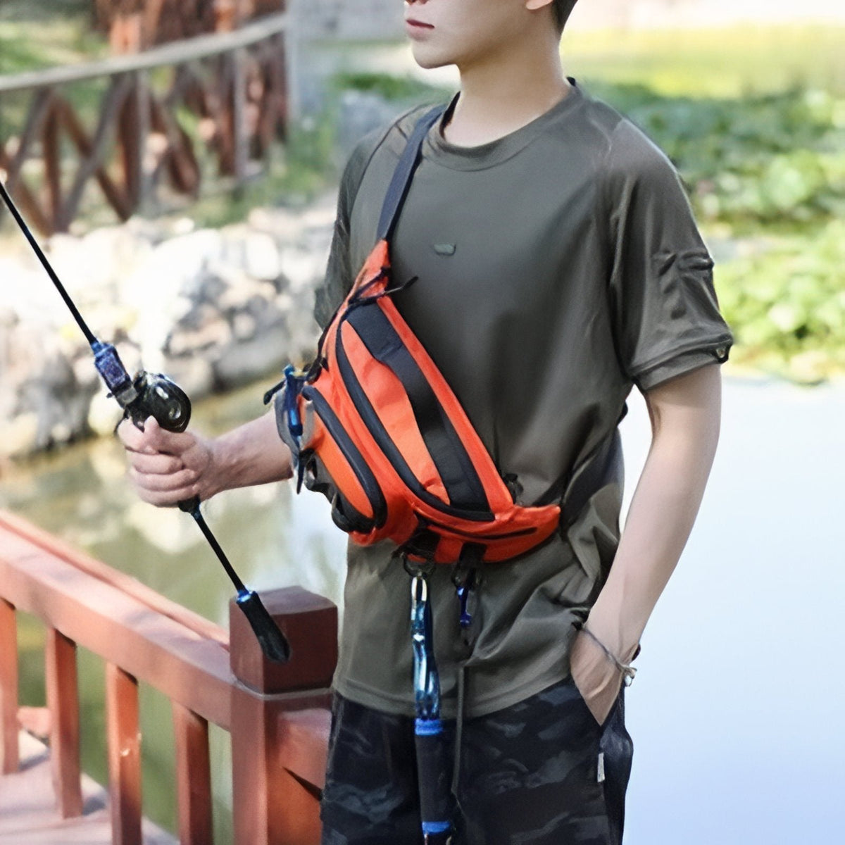 Angler wearing the orange hip pack across his chest by a lake, fishing rod in hand