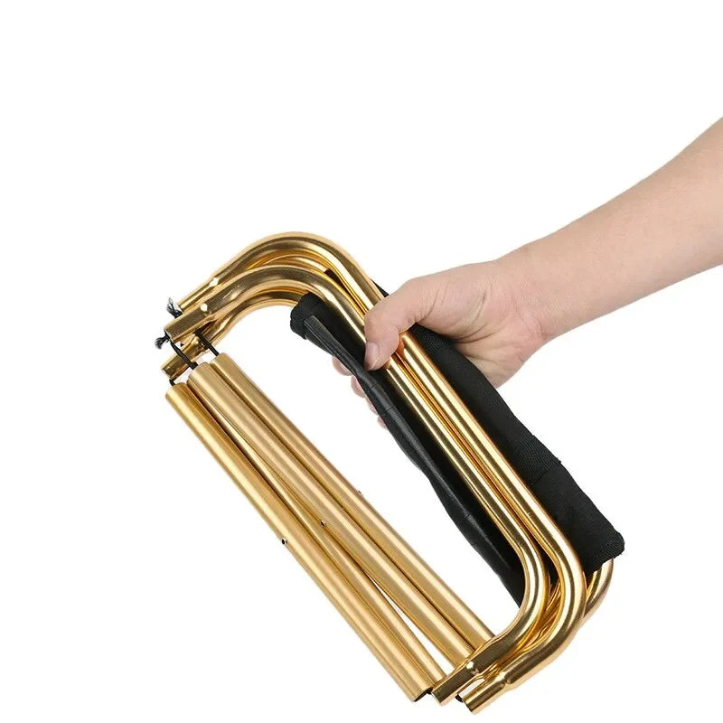 A hand holding the gold stool folded shut by its frame, showing how small the packed bundle is, against a white background.