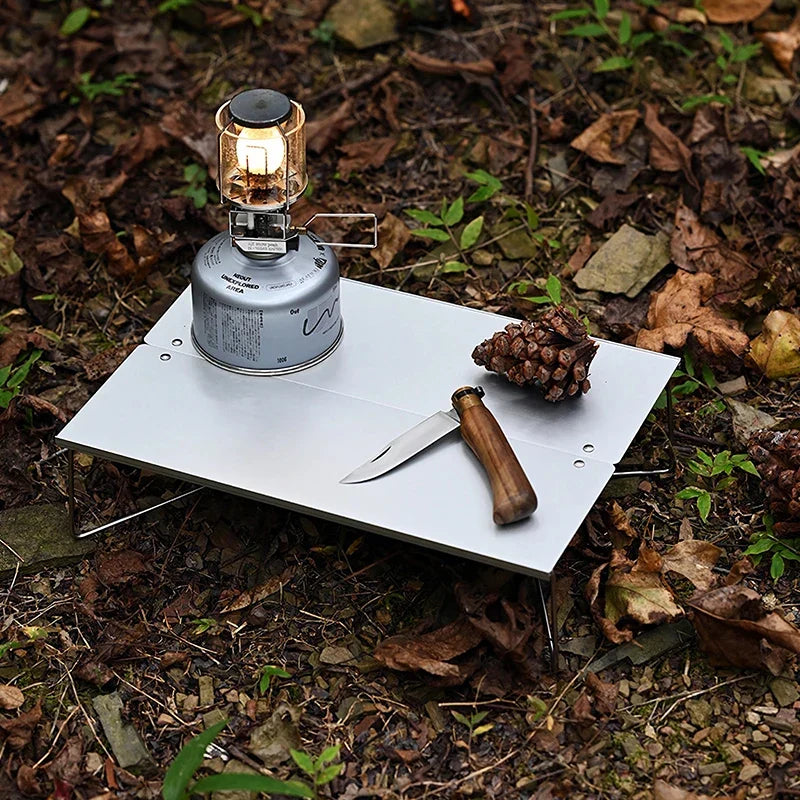 The low table on leaf litter with a lit gas lantern on a canister, a pine cone and a wooden-handled knife resting on the top.