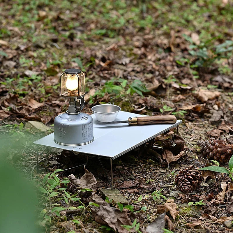 The table standing in leaf litter, holding a lit gas lantern on a fuel canister, a small steel bowl and a wooden-handled tool.