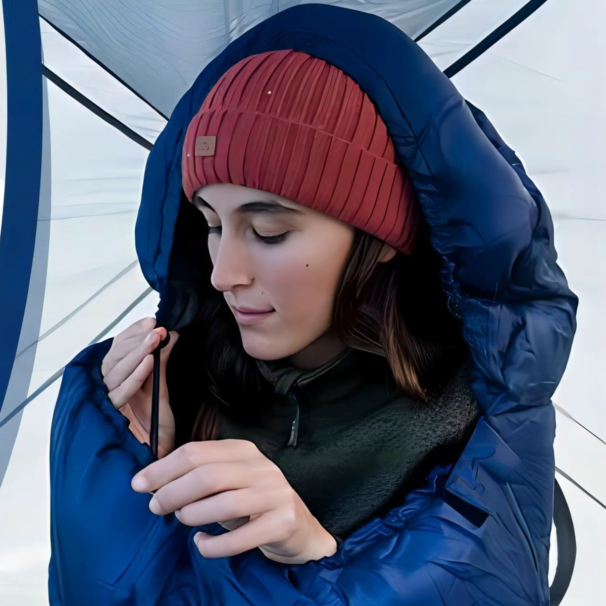 Woman in a red beanie zipping the navy mummy sleeping bag up to her chin inside a tent