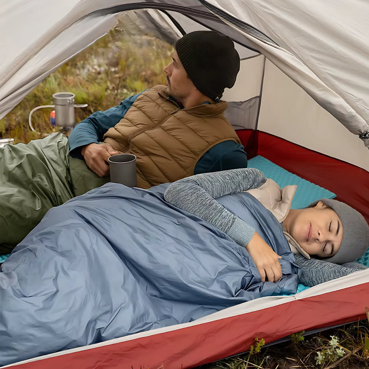 Woman asleep under a grey-blue envelope sleeping bag in a tent, a man in a green sleeping bag beside her holding a mug