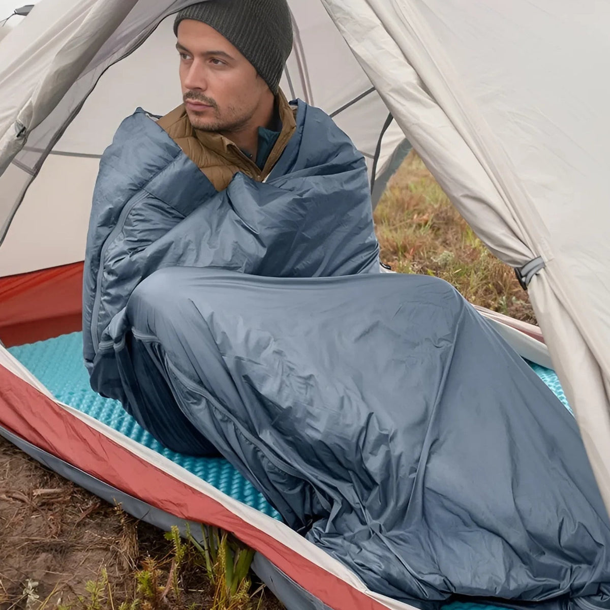 Man in a tent wrapped in an opened grey-blue envelope sleeping bag like a blanket