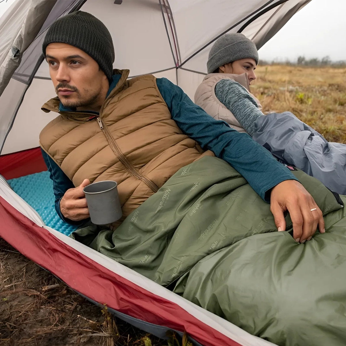 Man sitting up in a tent with a green envelope sleeping bag over his legs, holding a mug