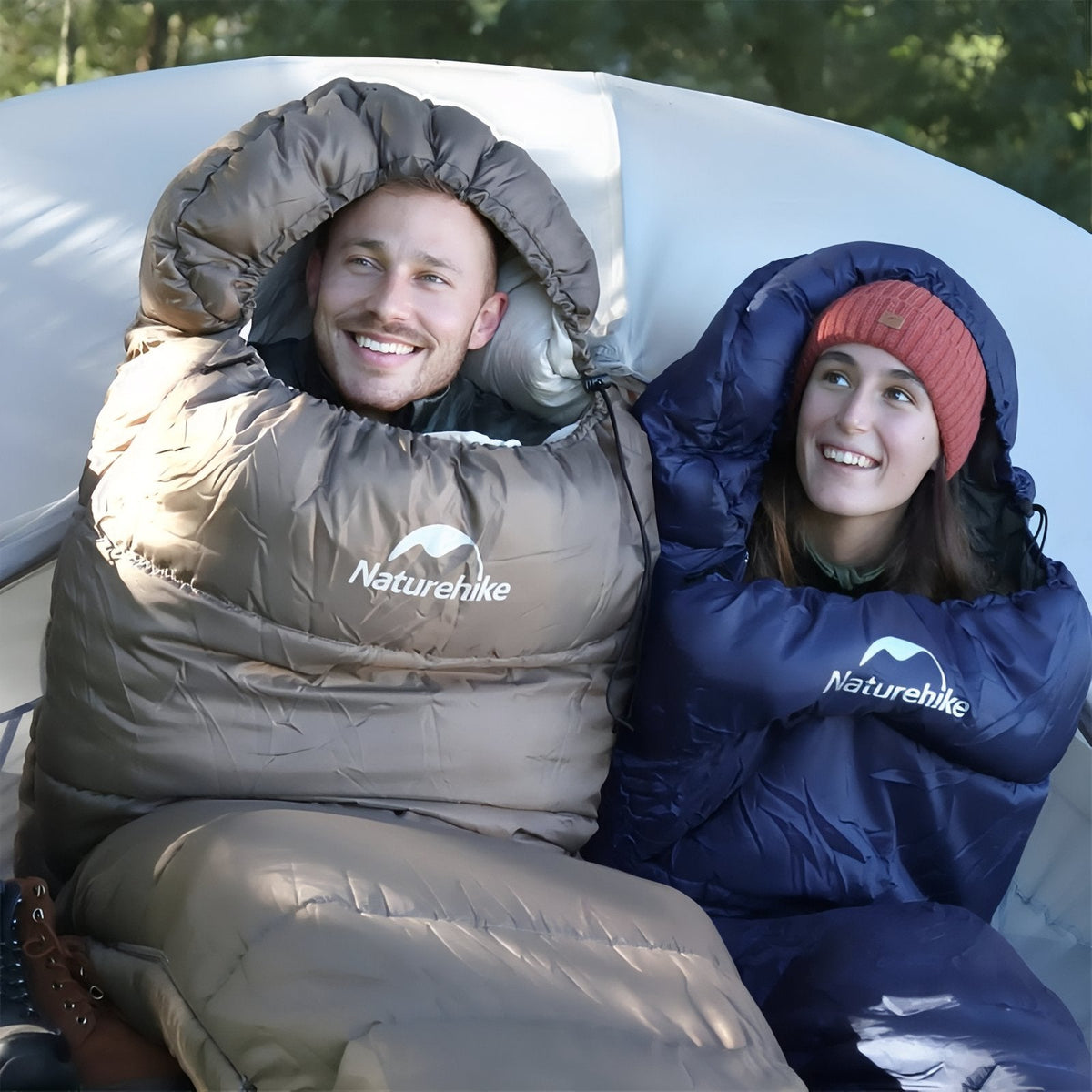 Two campers smiling in mummy sleeping bags in front of a tent, one khaki and one navy