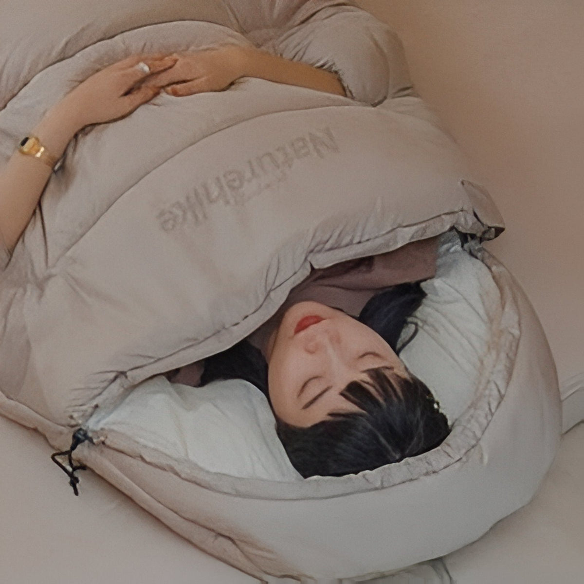 A woman lying in the light beige sleeping bag with the hood drawn around her face and the drawcord toggle at the side