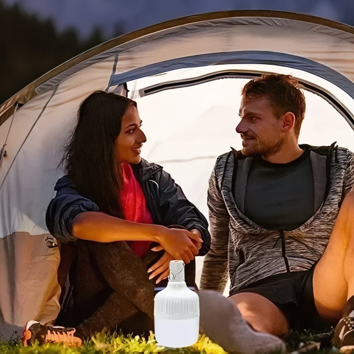 Couple sitting in a tent doorway at dusk with the LED bulb lantern glowing on the grass