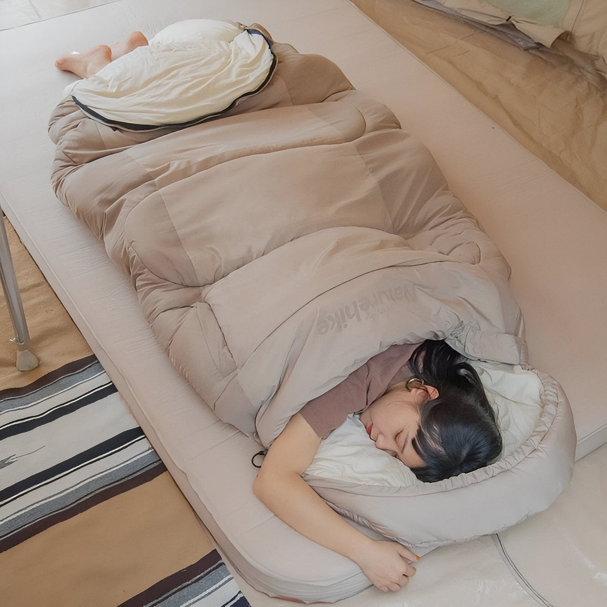 A woman asleep on her side in the light beige hooded sleeping bag on a camping mat, the hood open around her head
