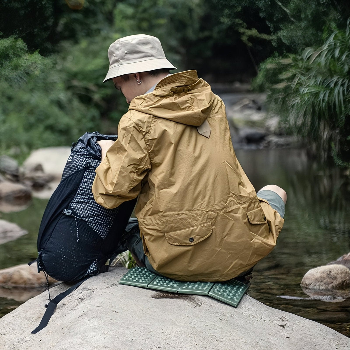 A hiker seen from behind sitting on the green sit pad on a rock by a river, reaching into his backpack