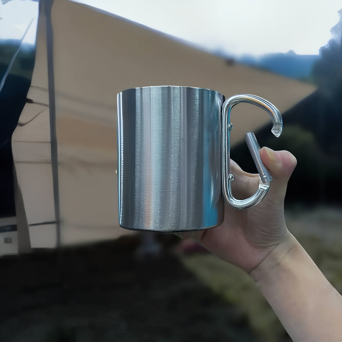 Hand holding the stainless steel mug by its carabiner handle in front of a tent