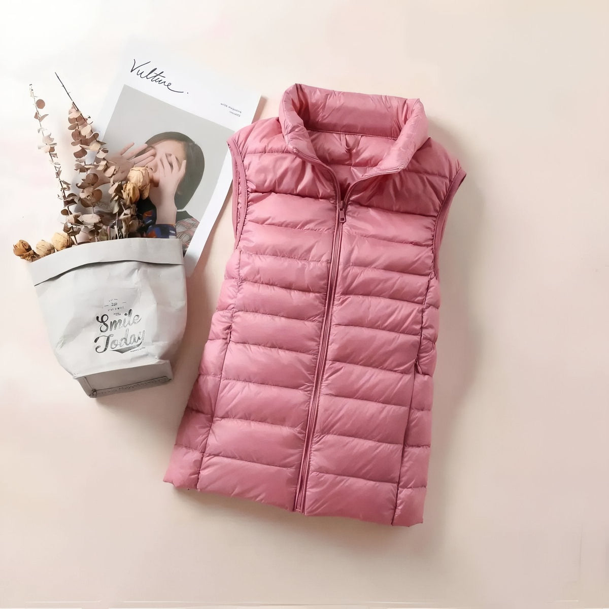 Powder pink quilted vest laid flat beside a magazine and dried flowers