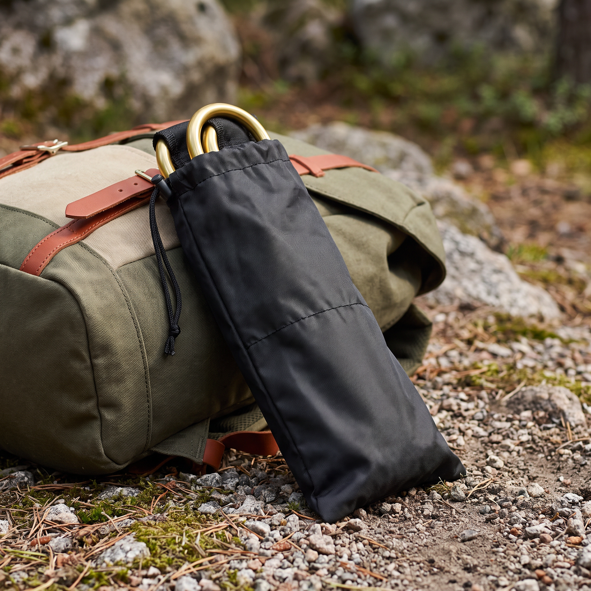 The stool folded shut inside its black drawstring pouch, leaning against a canvas rucksack on a gravel trail, with the gold tube bends showing above the drawcord.