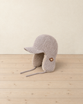 Beige knitted trapper hat with a long peak, plush-lined ear flaps, tie cords and a small leather patch, on a pale pine floor