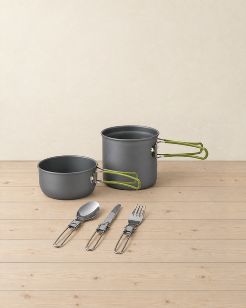 Solo camp cook set: a tall and a shallow grey aluminium pot with green-coated folding handles, and a folding spoon, knife and fork, on pale pine floorboards