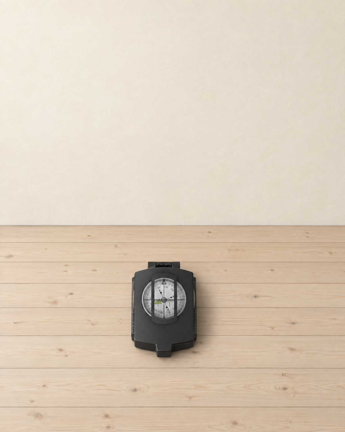 Closed black lensatic sighting compass with its white dial showing through the lid window, lying on a pale pine floor