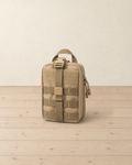 Khaki MOLLE pouch standing upright on a pale pine floor, with its empty hook-and-loop panel, front strap and buckle