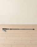 Black folding carbon and aluminium trekking pole lying extended on a pale pine floor, foam grip and wrist strap at the left, orange ring halfway down