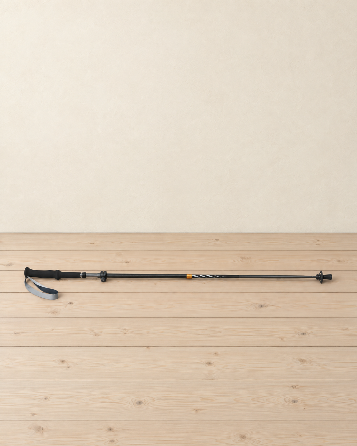 Black folding carbon and aluminium trekking pole lying extended on a pale pine floor, foam grip and wrist strap at the left, orange ring halfway down