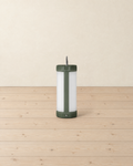 Cylindrical camping lantern with a white frosted diffuser and olive green caps, standing on pale pine floorboards against a cream wall