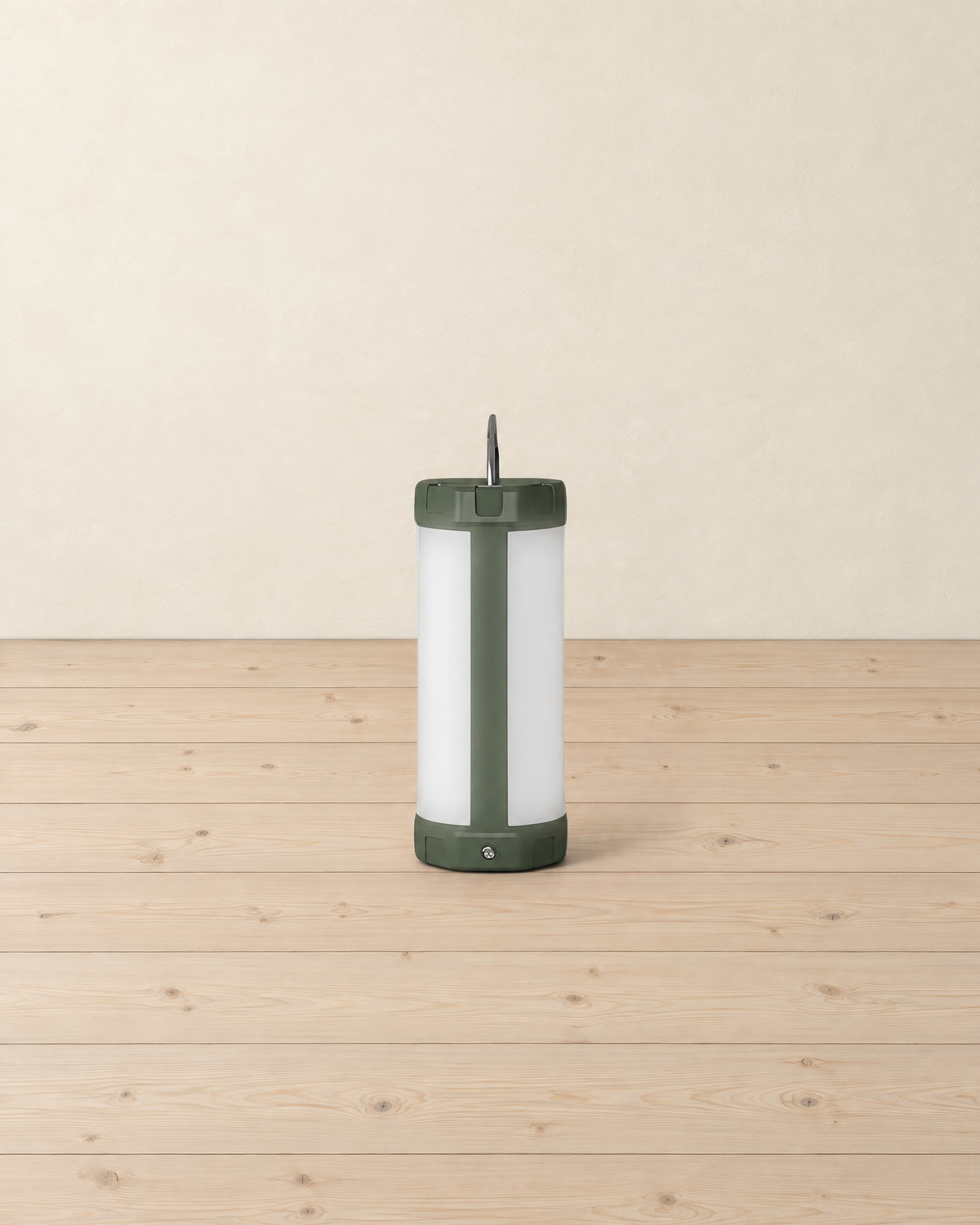Cylindrical camping lantern with a white frosted diffuser and olive green caps, standing on pale pine floorboards against a cream wall