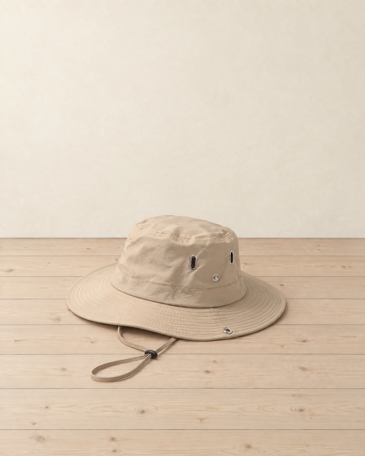 Beige wide-brim hiking hat with metal-ringed air vents and a chin cord with a black toggle, on pale pine floorboards