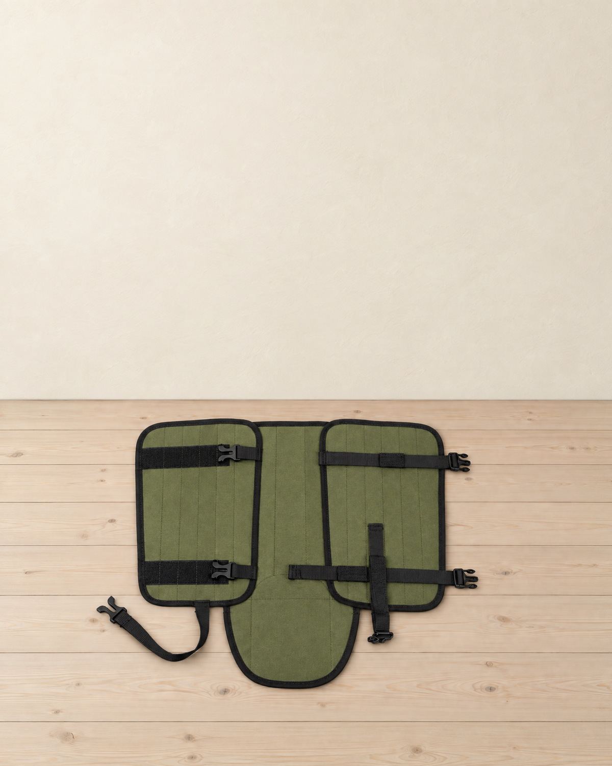 Army green hiking gaiter opened out flat on a pale pine floor, showing its hook-and-loop bands, two buckle straps, vertical strap and under-boot strap