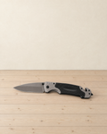 Open folding knife with a grey drop-point blade and a black and grey handle lying on a pale pine floor