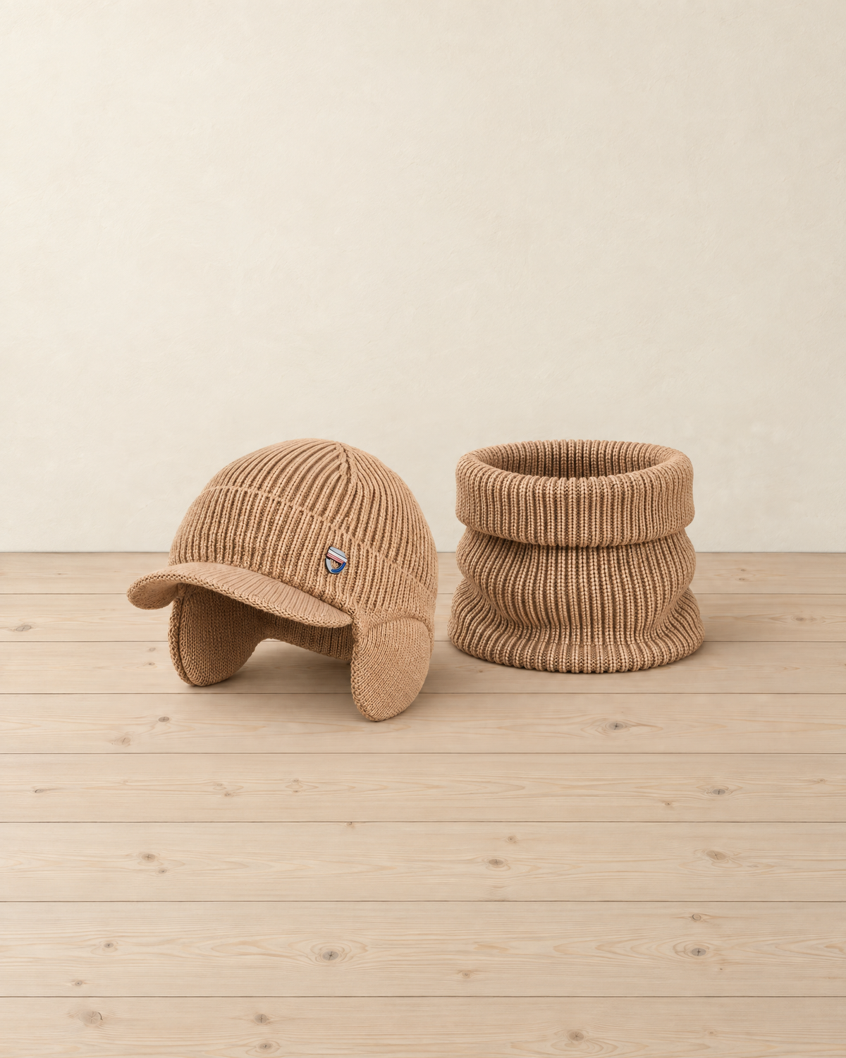 Camel rib-knit winter hat with a short peak, ear flaps and a small metal badge, beside its matching neck warmer on a pale pine floor