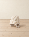 Cream ribbed knit beanie with a turn-up cuff and ear flaps knitted with a brown snowflake, on a pale pine floor