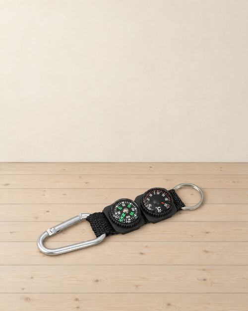 Black webbing keyring with a small compass and a Celsius dial thermometer, a silver carabiner and a split ring, lying on pale pine floorboards