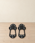 Pair of black rubber ice cleats lying stud side up on a pale pine floor, each with 11 metal studs, 7 under the forefoot and 4 under the heel