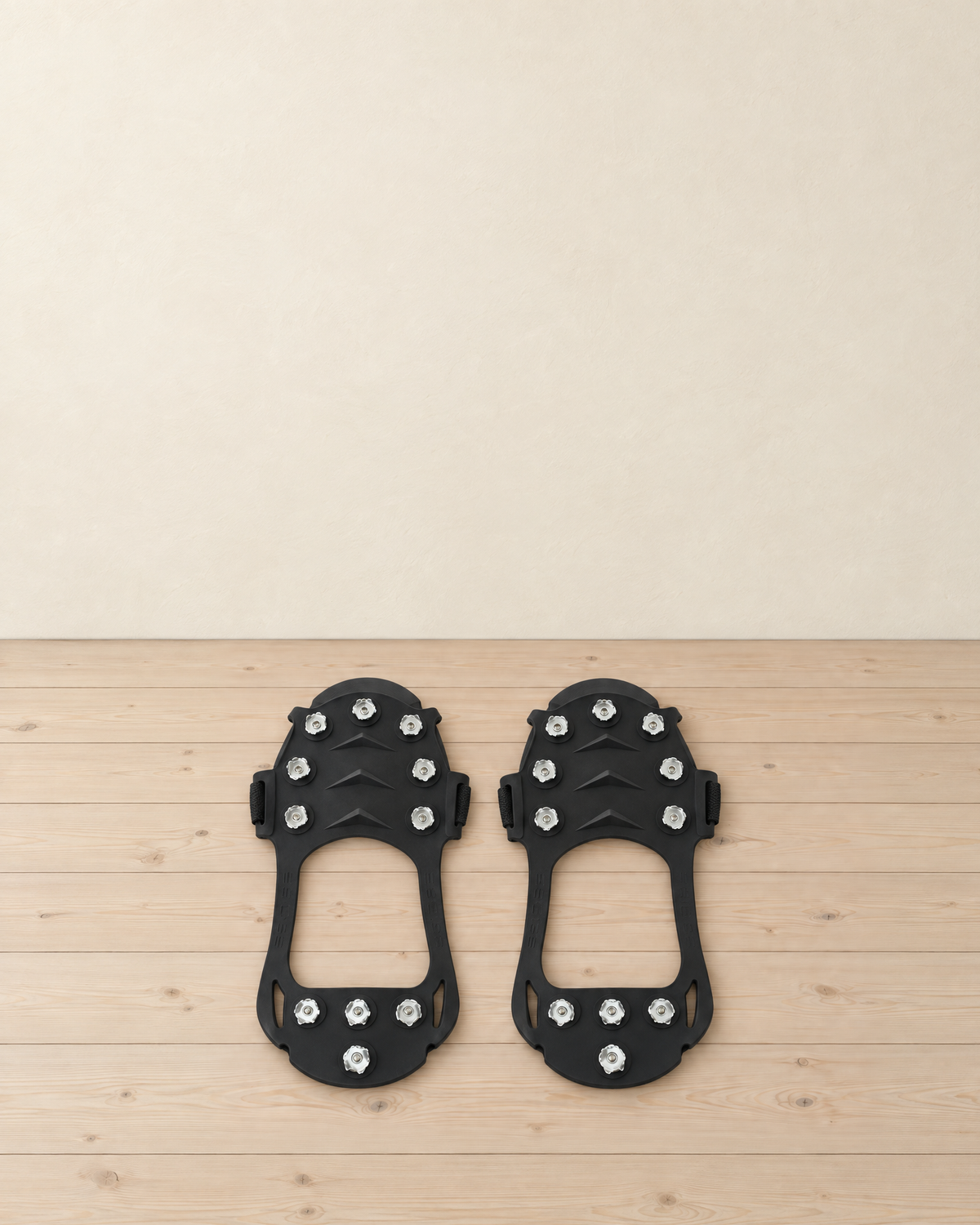 Pair of black rubber ice cleats lying stud side up on a pale pine floor, each with 11 metal studs, 7 under the forefoot and 4 under the heel