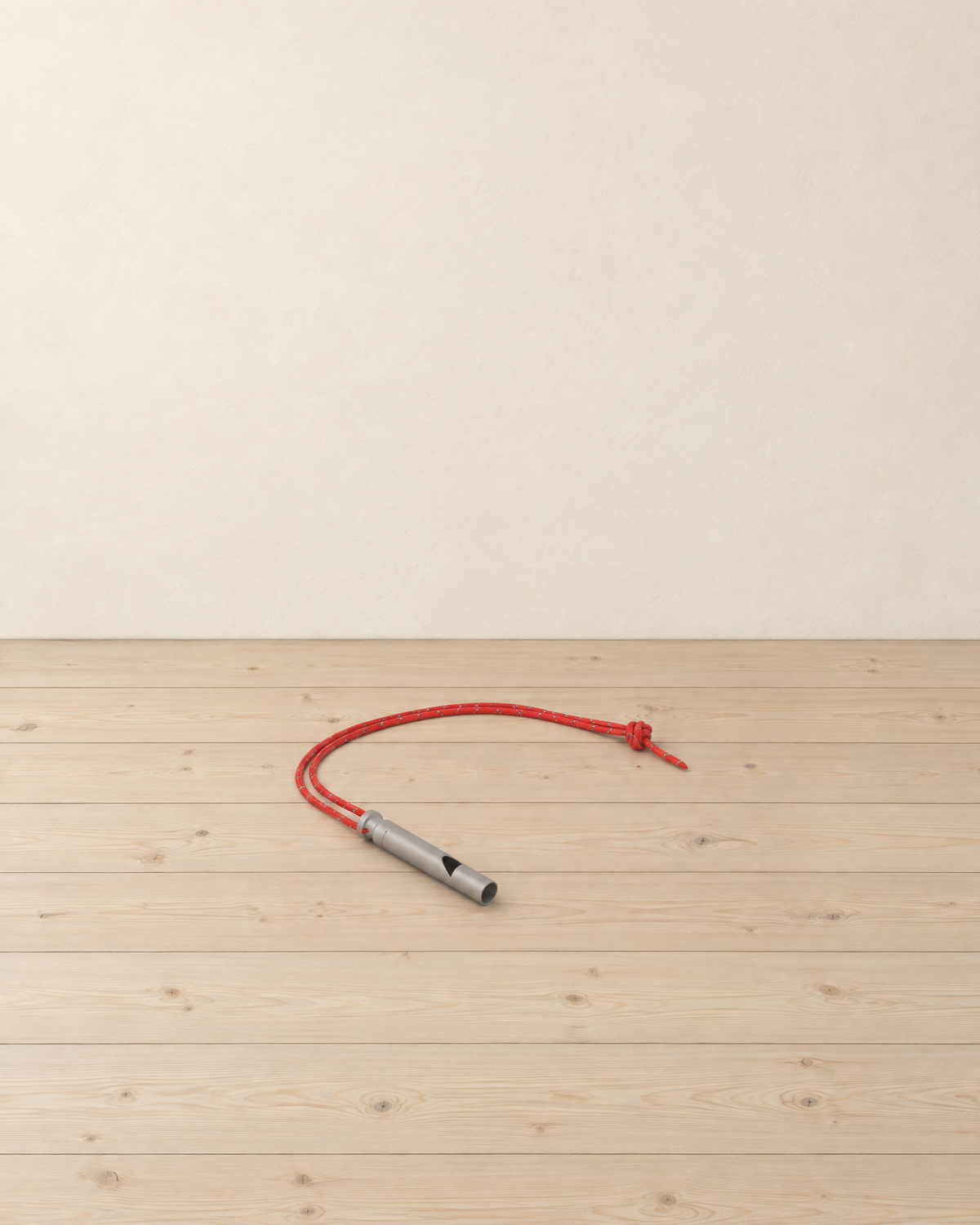 Titanium Grey whistle on its red reflective cord lying on a pale pine floor against a cream wall