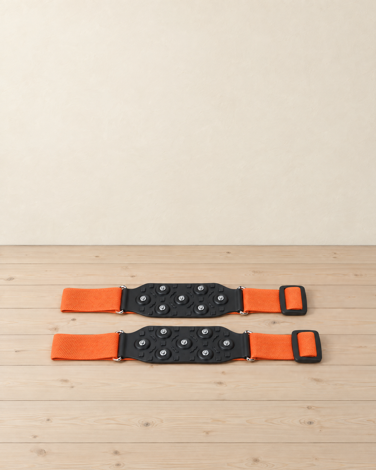 Pair of mid-sole ice cleats lying flat: black rubber pads with seven steel studs each and orange elastic straps with black buckles